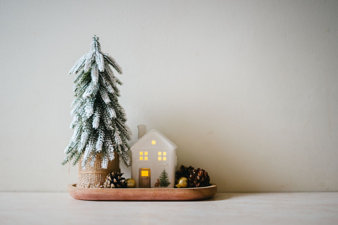 A minimalist Christmas scene with a small snowy tree, lit house, and pinecones on a tray indoors.